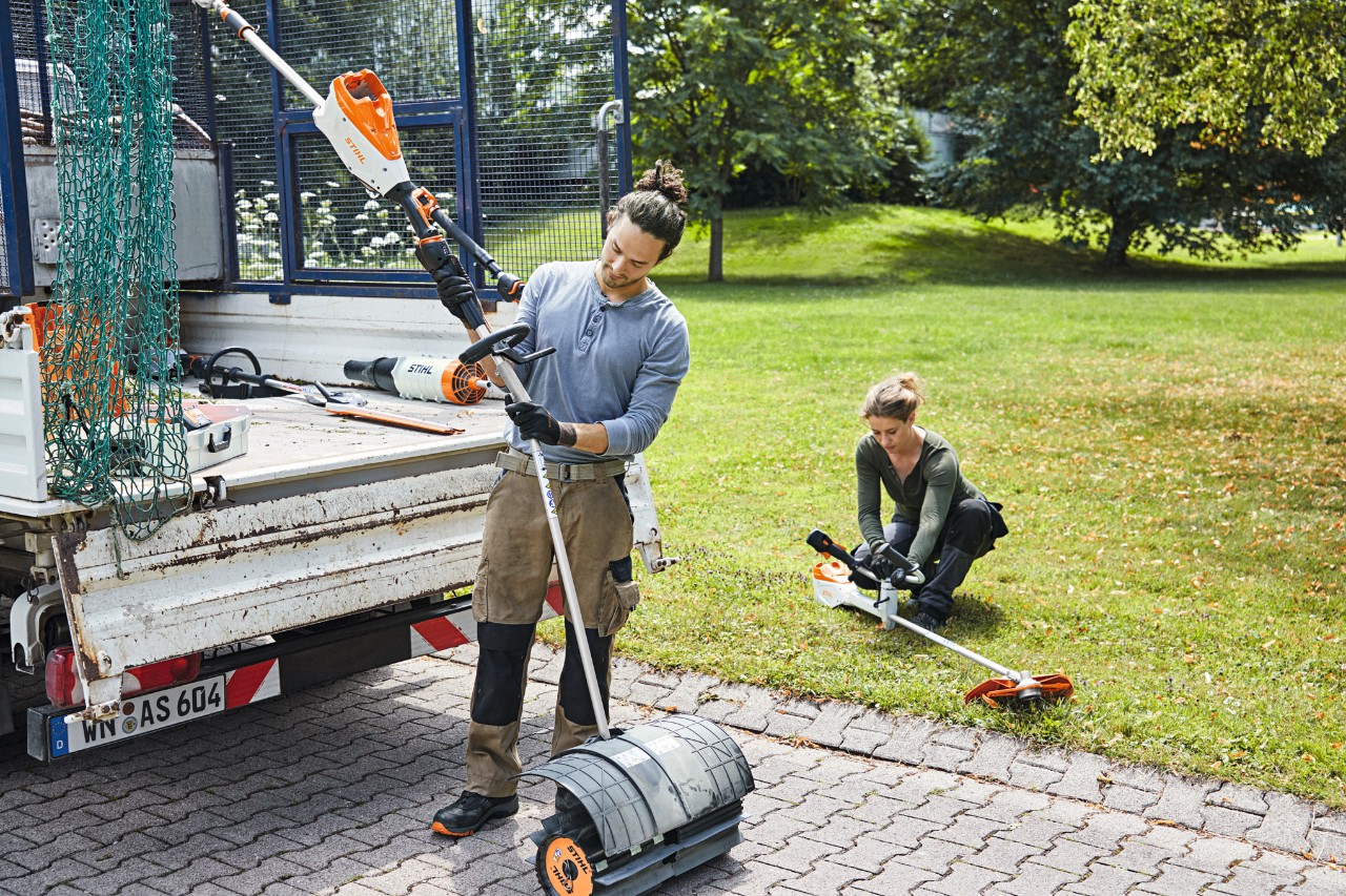 Muškarac i žena rukuje STIHL bateriijskim uređajima pored vozila sa otvorenom utovarnom površinom, u pozadini se vidi livada