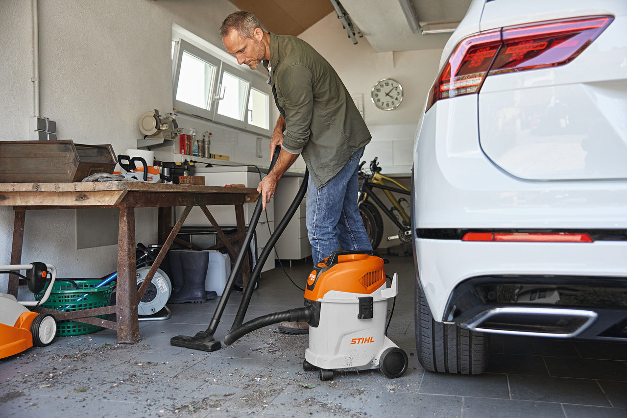Muškarac usisava pod garaže koristeći vlažno/suvi usisivač, pored njega radni sto i beli automobil
